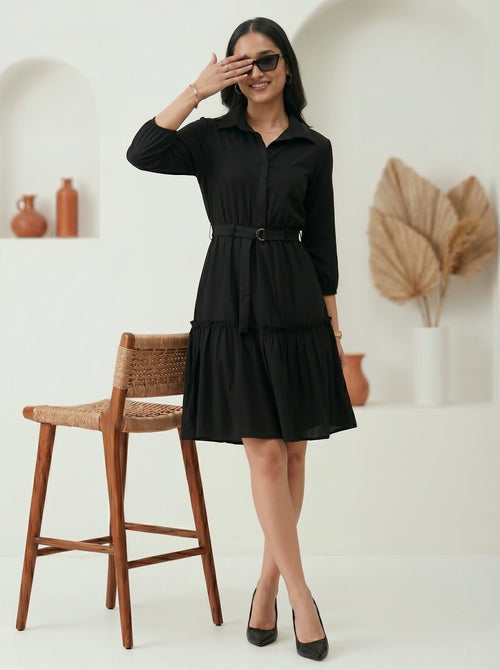 Woman wearing a black dress standing in a room with a wooden chair and decorative items.