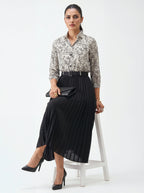 Woman in a patterned blouse and black skirt sitting on a white stool against a white background