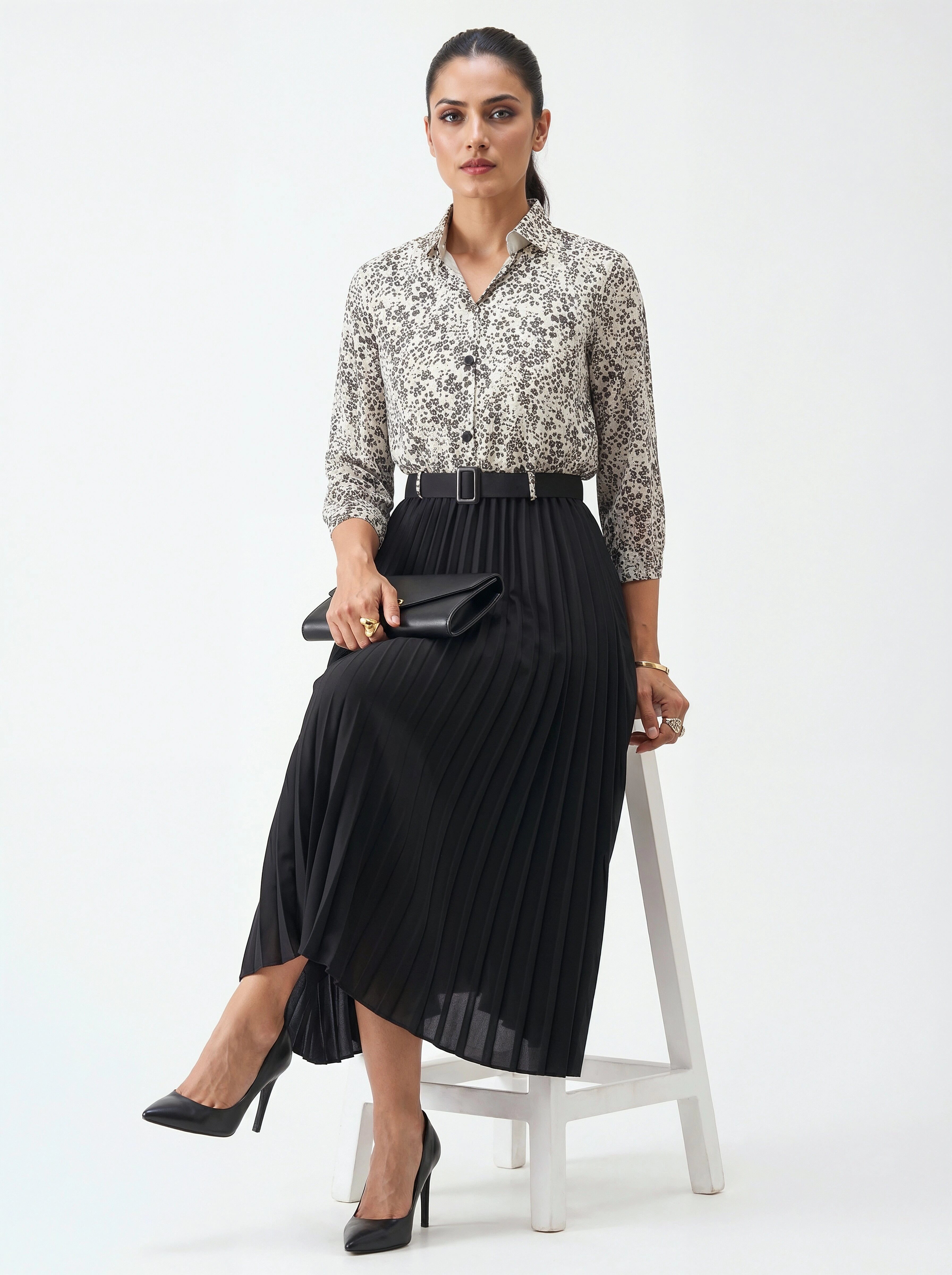 Woman in a patterned blouse and black skirt sitting on a white stool against a white background