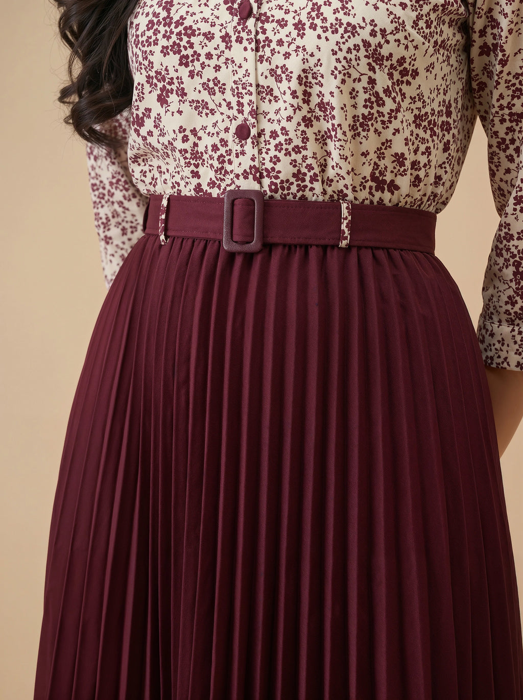 Woman wearing a floral blouse and burgundy pleated skirt with a belt.