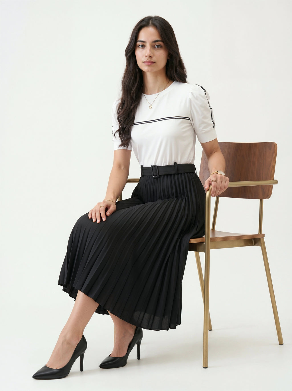 Woman sitting on a chair wearing a white top and black pleated skirt against a white background