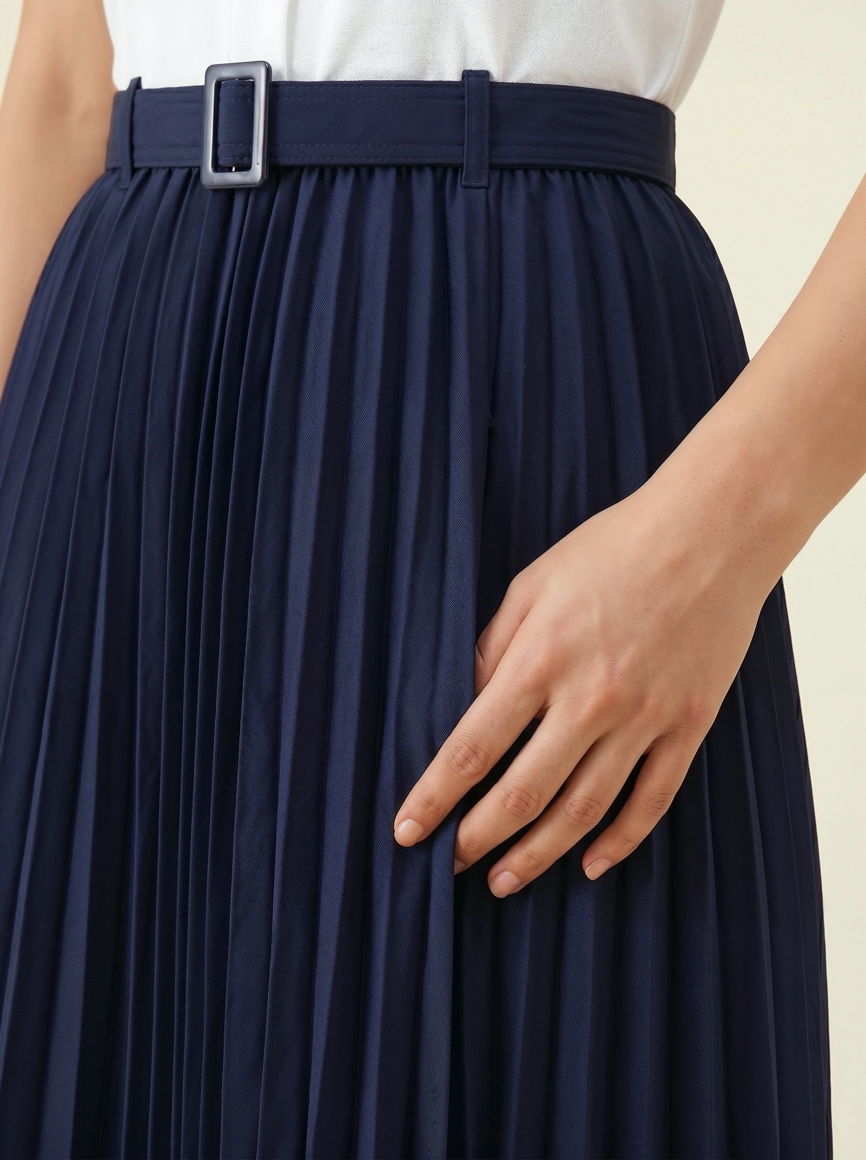 Navy pleated skirt with a belt and hand adjusting it on a neutral background