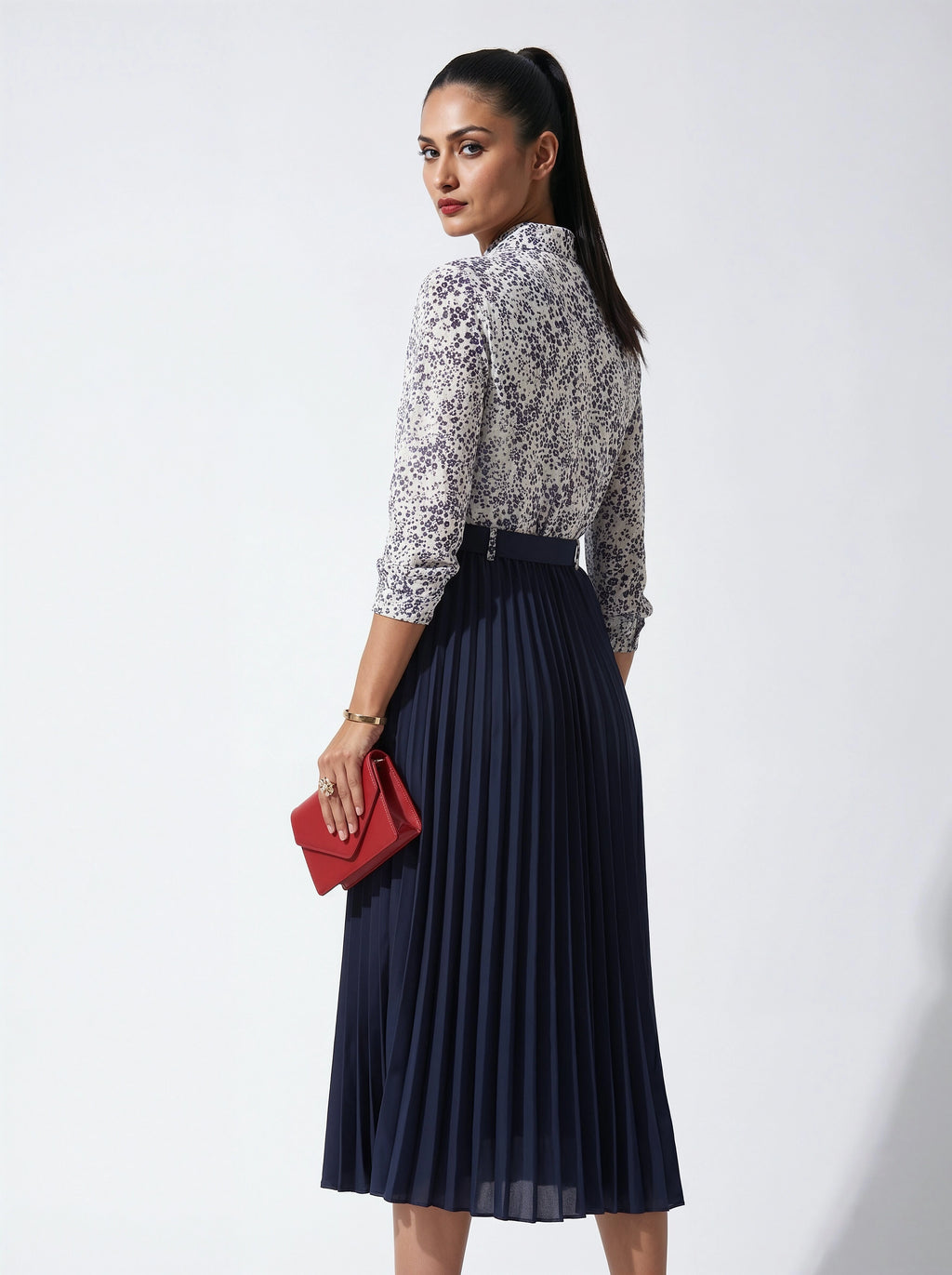 Woman wearing a patterned blouse and navy pleated skirt holding a red clutch against a white background
