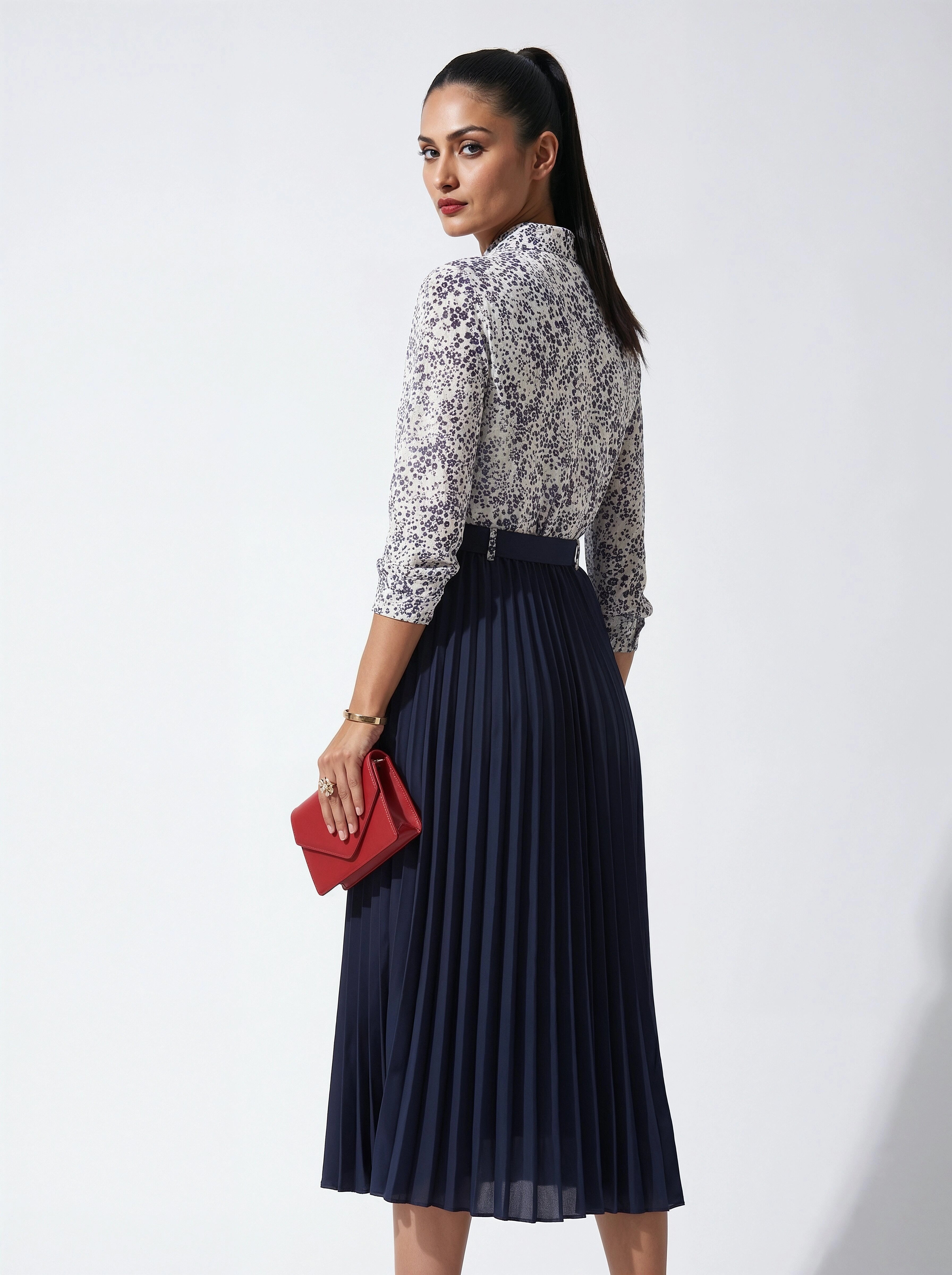 Woman wearing a patterned blouse and navy pleated skirt holding a red clutch against a white background