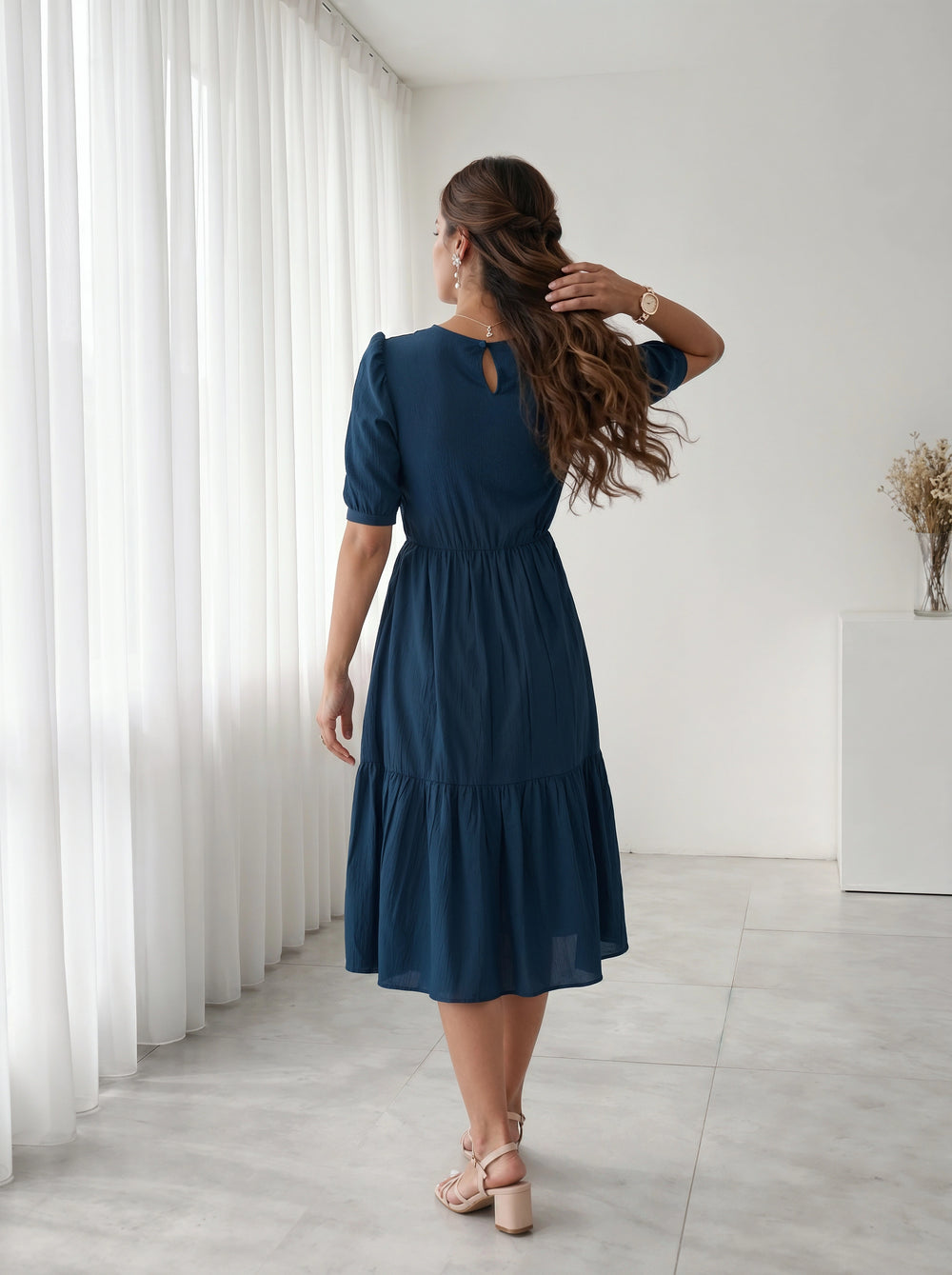 Woman in a blue dress standing in a room with white curtains and a white cabinet.