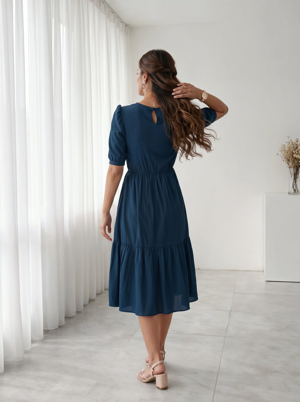 Woman in a blue dress standing in a room with white curtains and a white cabinet.