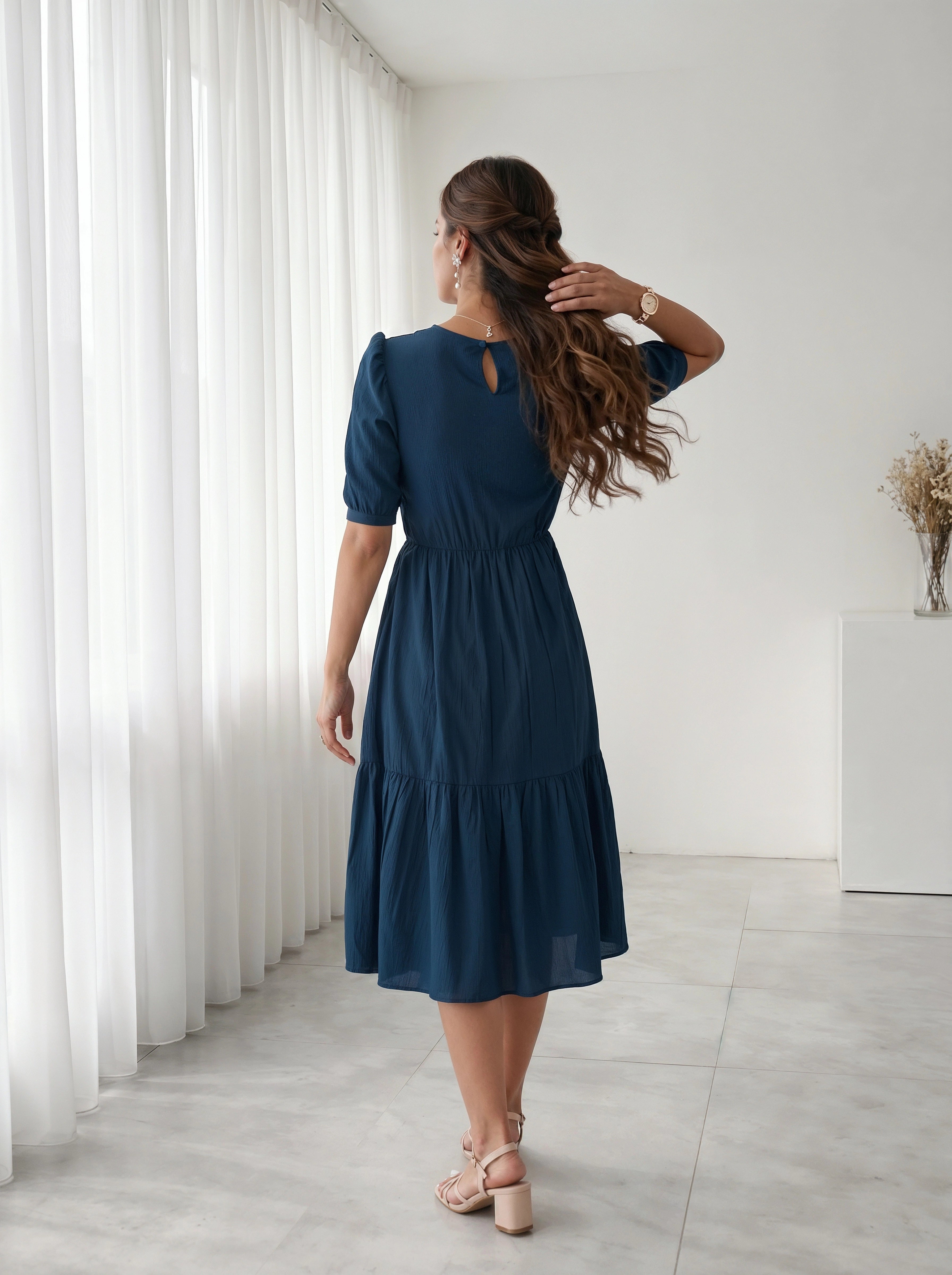 Woman in a blue dress standing in a room with white curtains and a white cabinet.