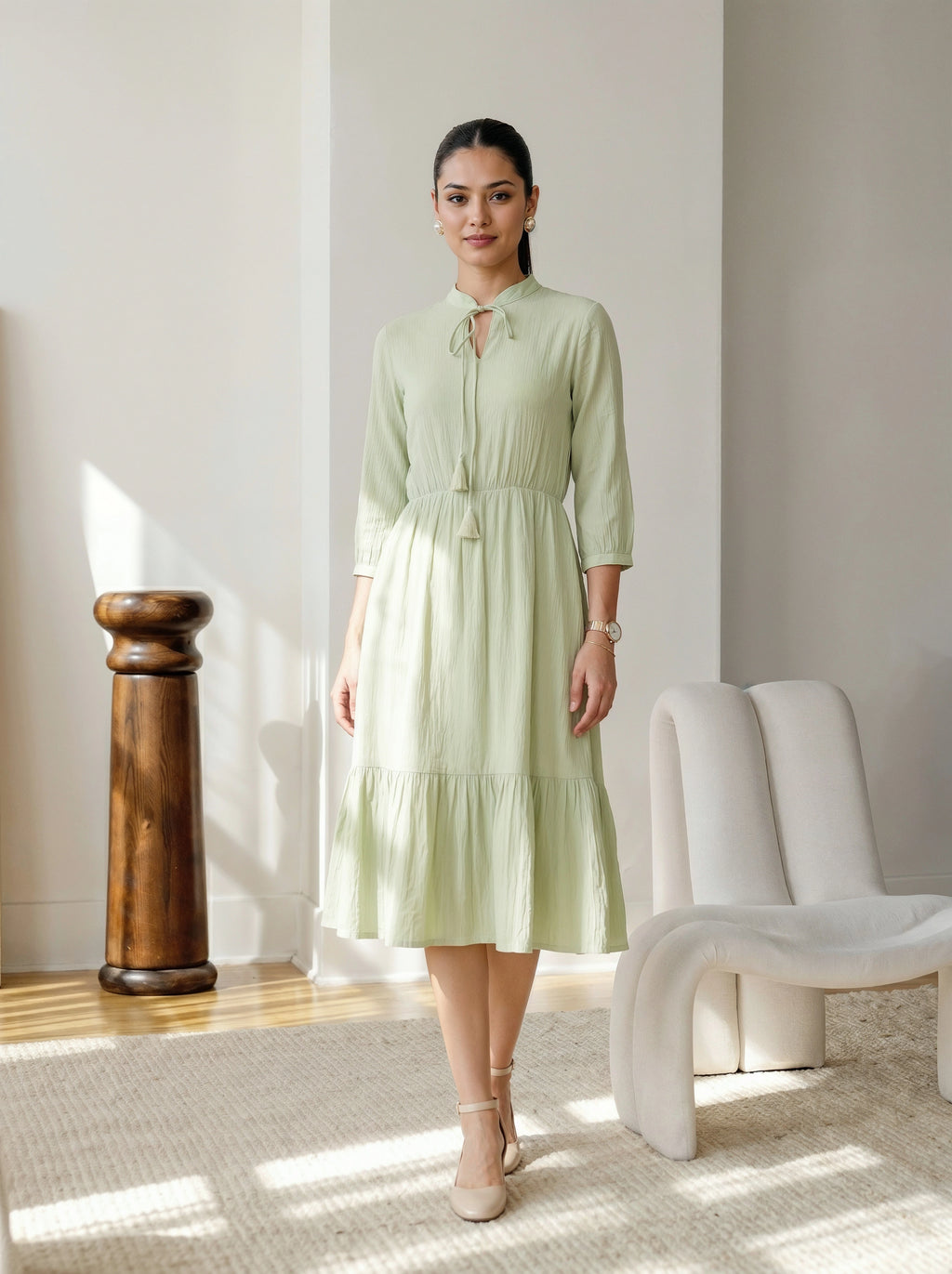 Woman in a light green dress standing in a well-lit room with a wooden column and white chair.