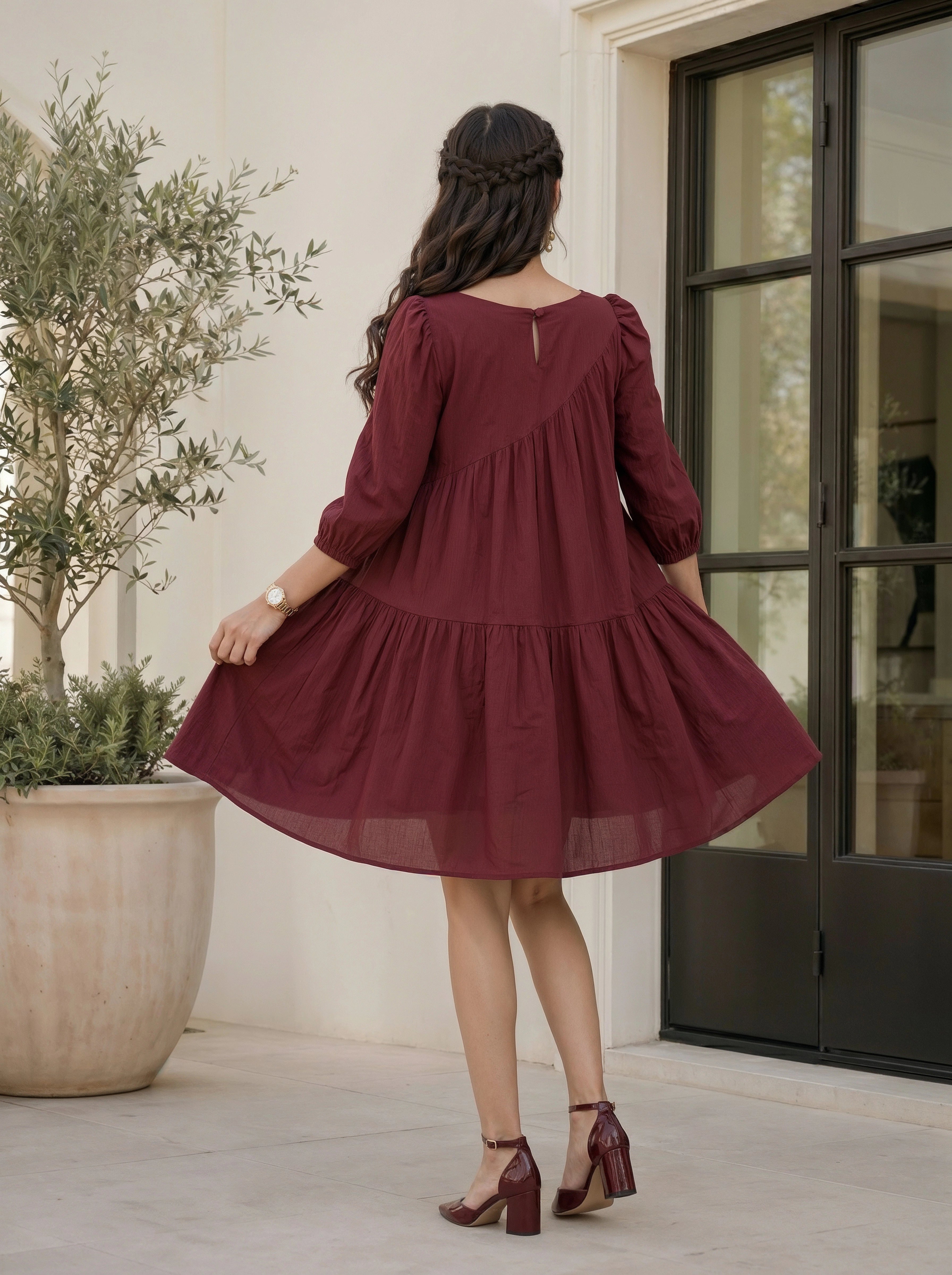 Woman in a burgundy dress standing in front of a door with a plant to her left.