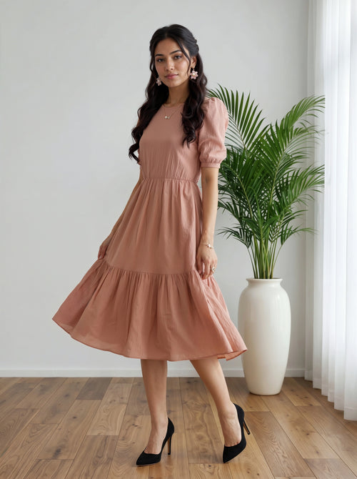 Woman in a pink dress standing in a room with a plant and white curtain.