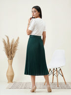 Woman wearing a white top and green pleated skirt standing in a minimalistic room with a chair and vase.