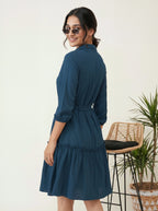 Woman wearing a blue dress standing in a room with a chair and plant.