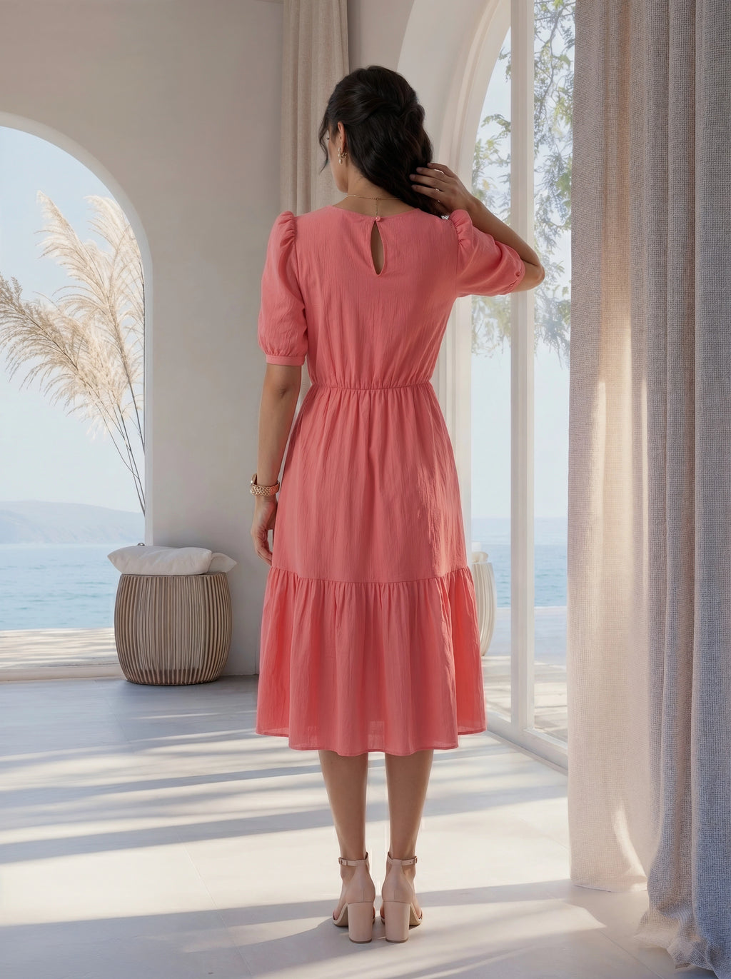 Woman in a pink dress standing in a bright room with large windows and a view of the ocean.