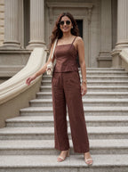 A woman stands on stone steps in front of a classical building with columns, wearing a rust-brown sleeveless camisole top and matching wide-leg pants. She is carrying a cream shoulder bag and wearing beige heeled sandals.