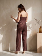 A woman stands with her back to the camera wearing a brown sleeveless jumpsuit with thin straps, a structured waist, and wide legs, paired with beige high heels in a sunlit room.