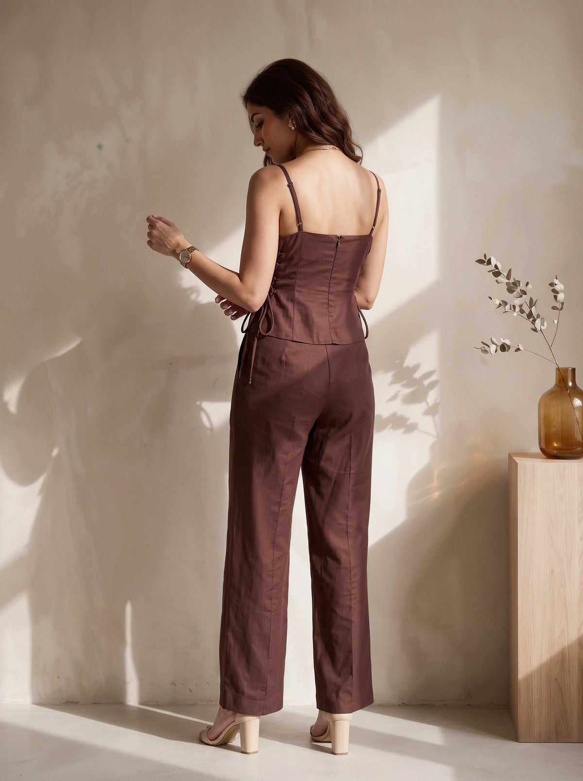 A woman stands with her back to the camera wearing a brown sleeveless jumpsuit with thin straps, a structured waist, and wide legs, paired with beige high heels in a sunlit room.