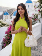 A woman wearing a bright lime green halter-neck dress stands outdoors in a Santorini-style setting with white buildings, blue domes, and pink flowers. She is holding a cream-colored handbag with pearl-like handles.