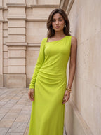 A woman wearing a bright lime green long-sleeve maxi dress with an asymmetrical neckline, posing against a beige stone building wall.