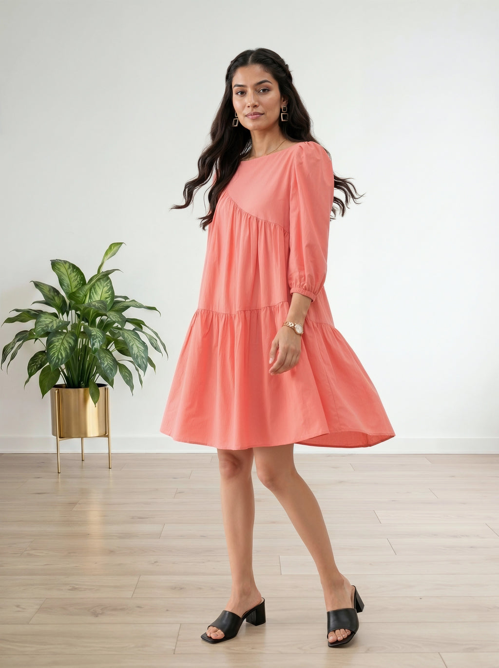 Woman wearing a coral dress standing in a room with a plant and white wall.