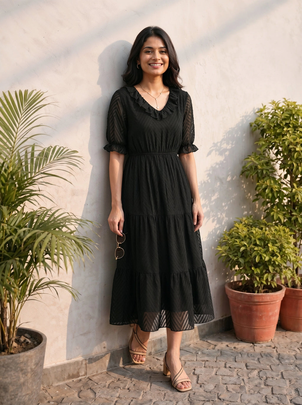 Woman in a black dress standing against a light wall with plants around.