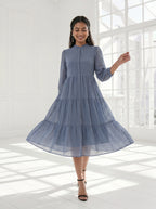 Woman wearing a blue dress standing in a bright room with large windows.
