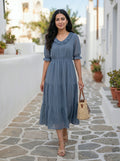 Woman in a blue dress standing on a stone path with plants and buildings in the background