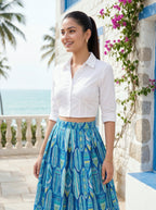 Woman wearing a white shirt and blue patterned skirt standing in front of a stone building with flowers.