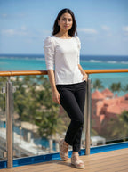 Woman standing on a balcony with ocean view wearing a white blouse and black pants.