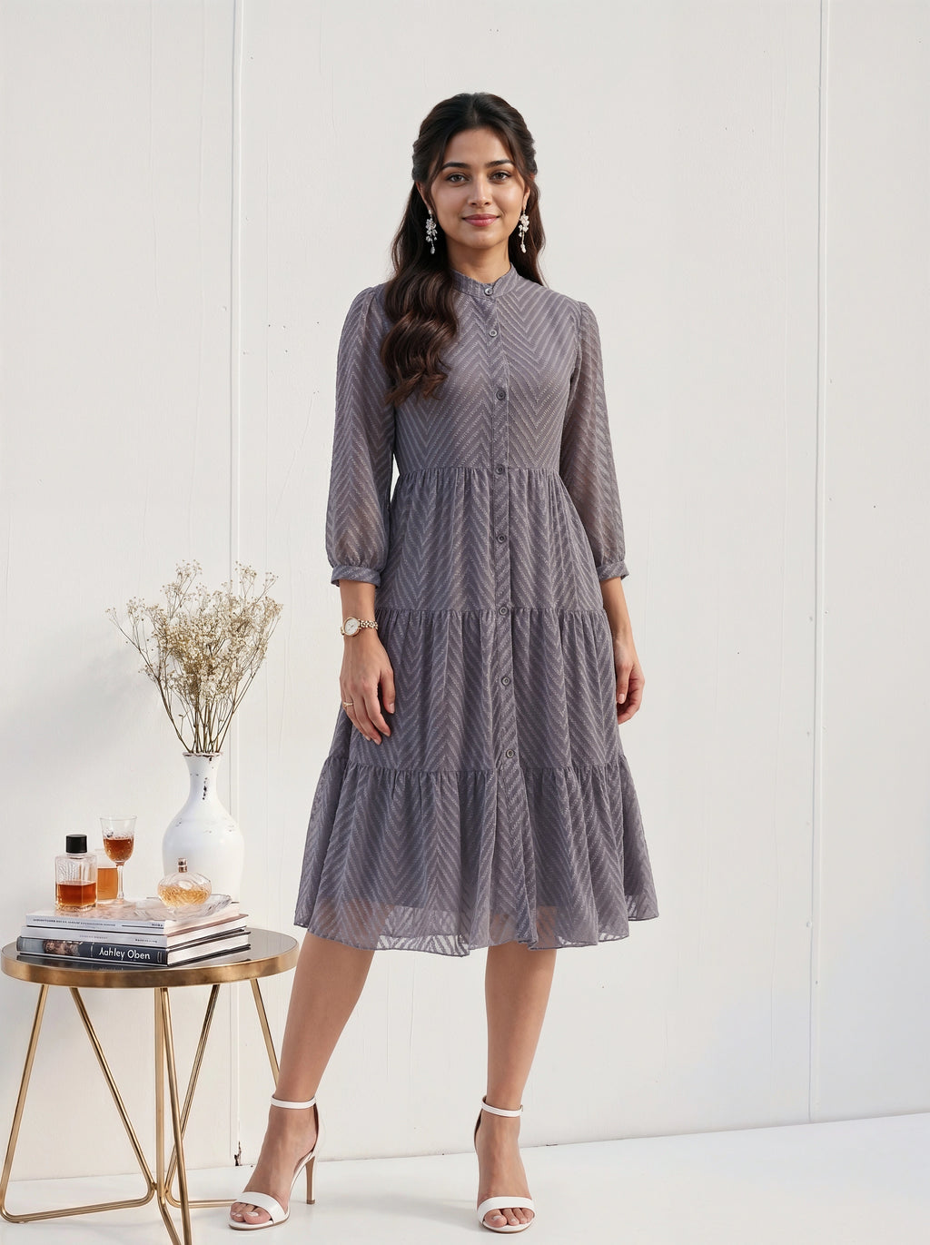 Woman wearing a gray patterned dress standing next to a small table with decor items on a white background