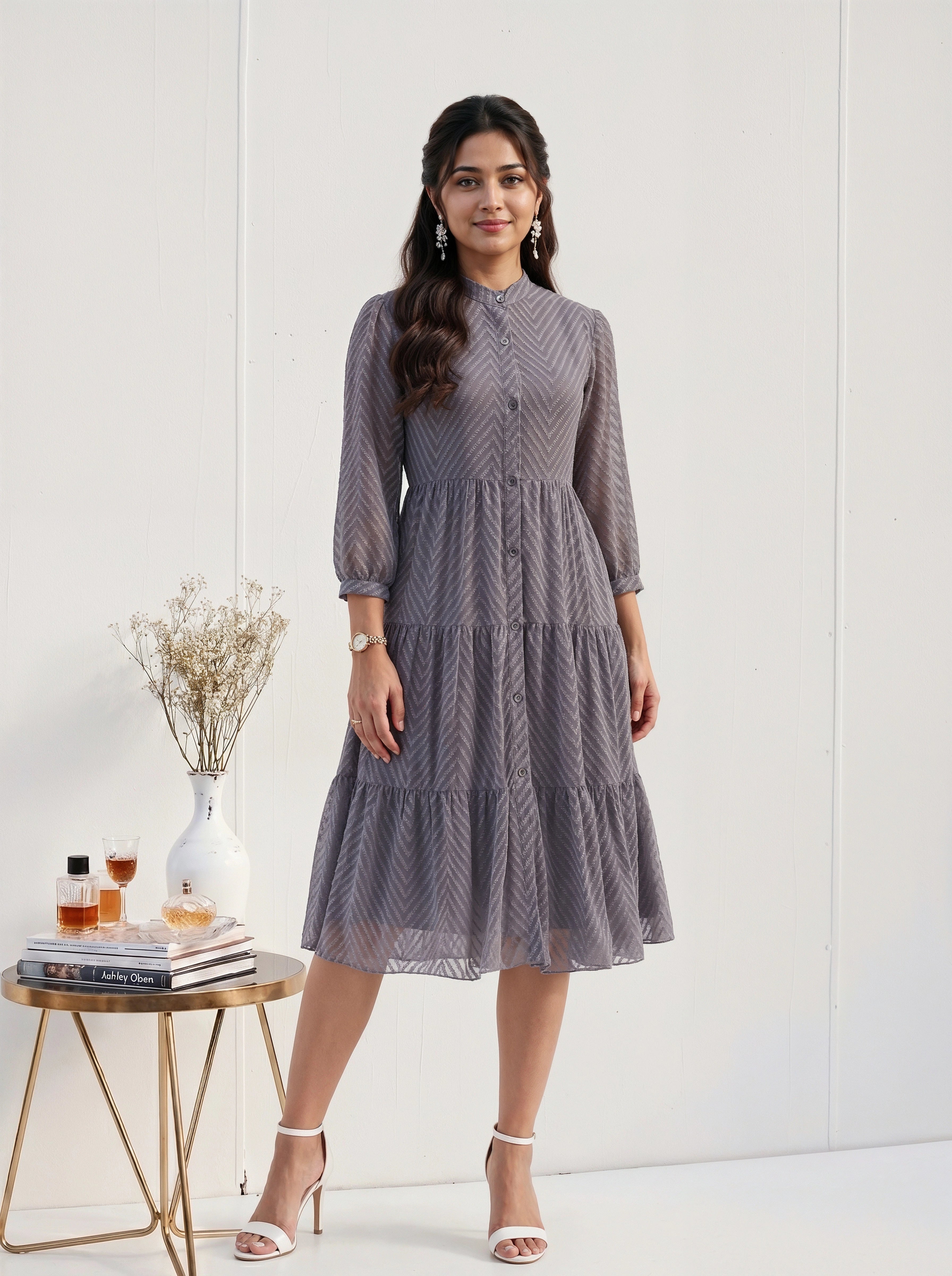 Woman wearing a gray patterned dress standing next to a small table with decor items on a white background