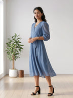 Woman in a blue dress standing in a room with a plant and wooden stool.