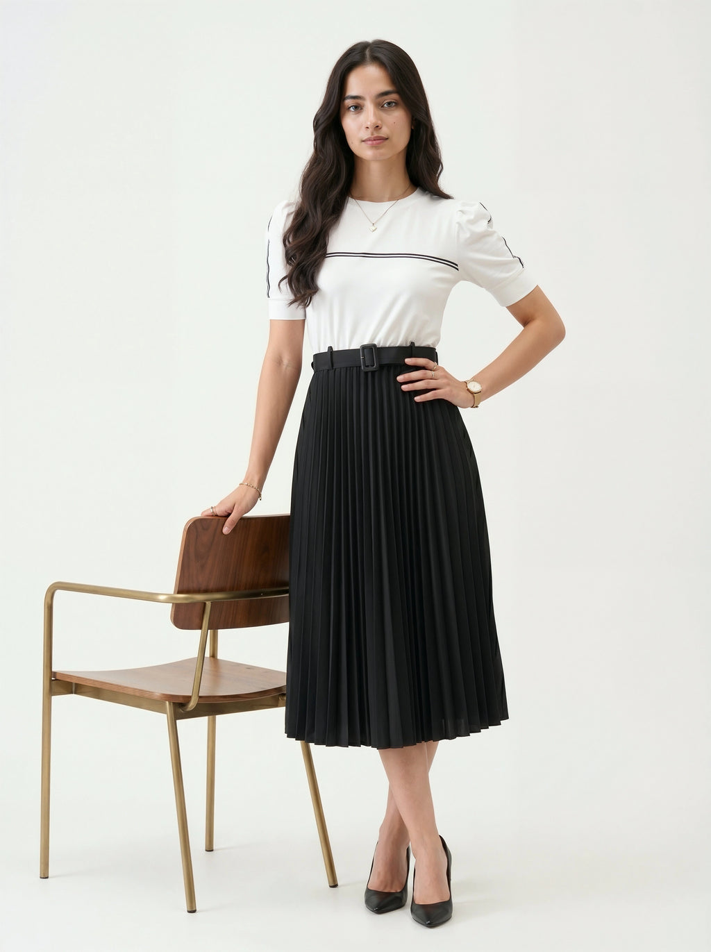 Woman wearing a white top and black pleated skirt standing next to a chair on a white background