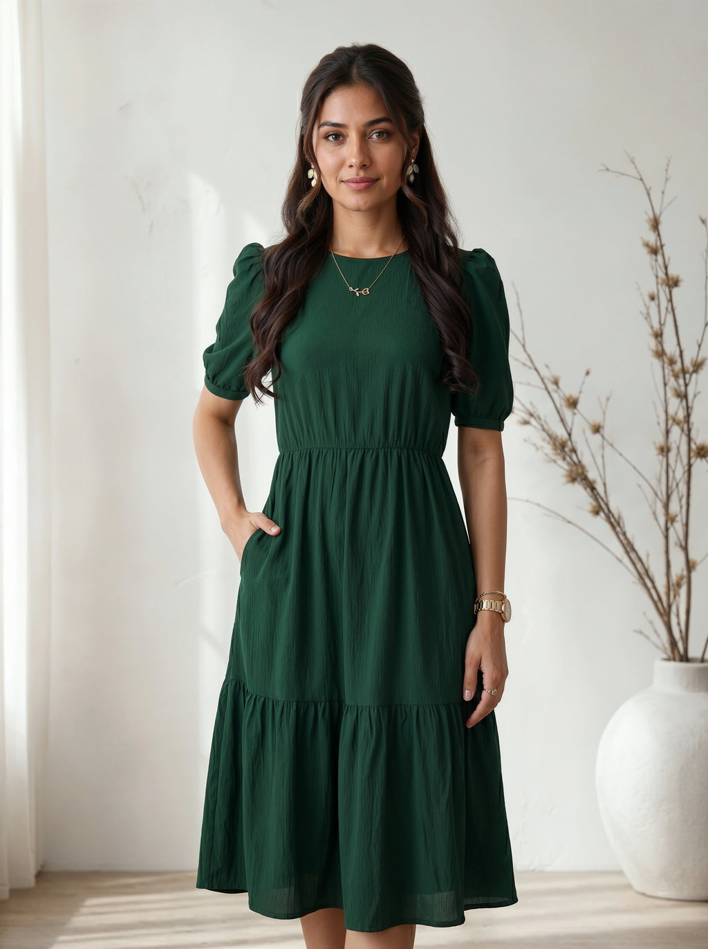 Woman wearing a green dress standing in a room with a white curtain and vase.
