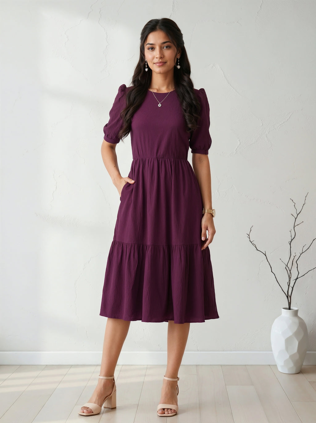 Woman wearing a purple dress standing against a white wall with a vase in the background.