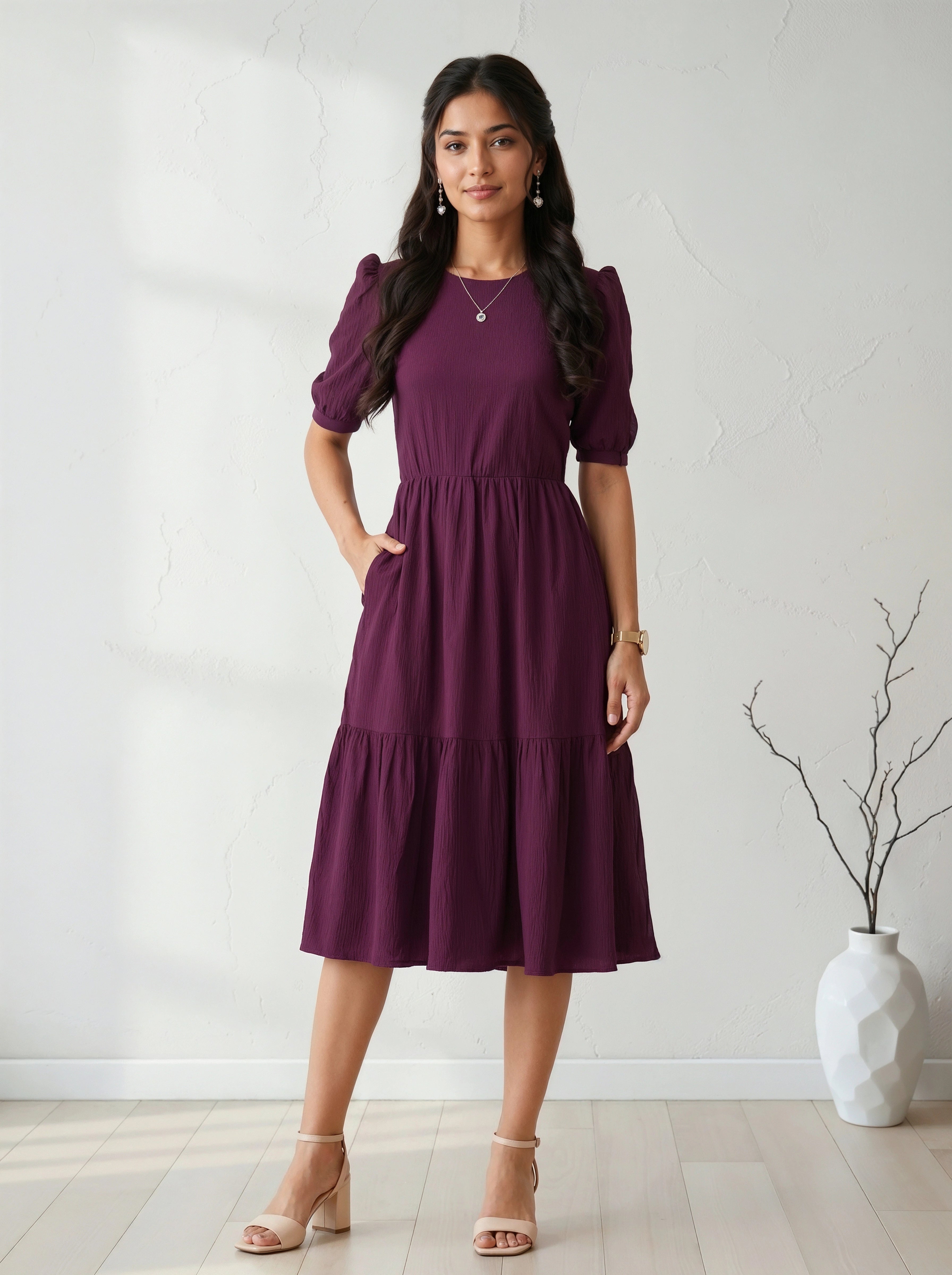 Woman wearing a purple dress standing against a white wall with a vase in the background.