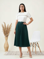 Woman wearing a white top and green pleated skirt standing in a minimalistic room with a chair and plant.