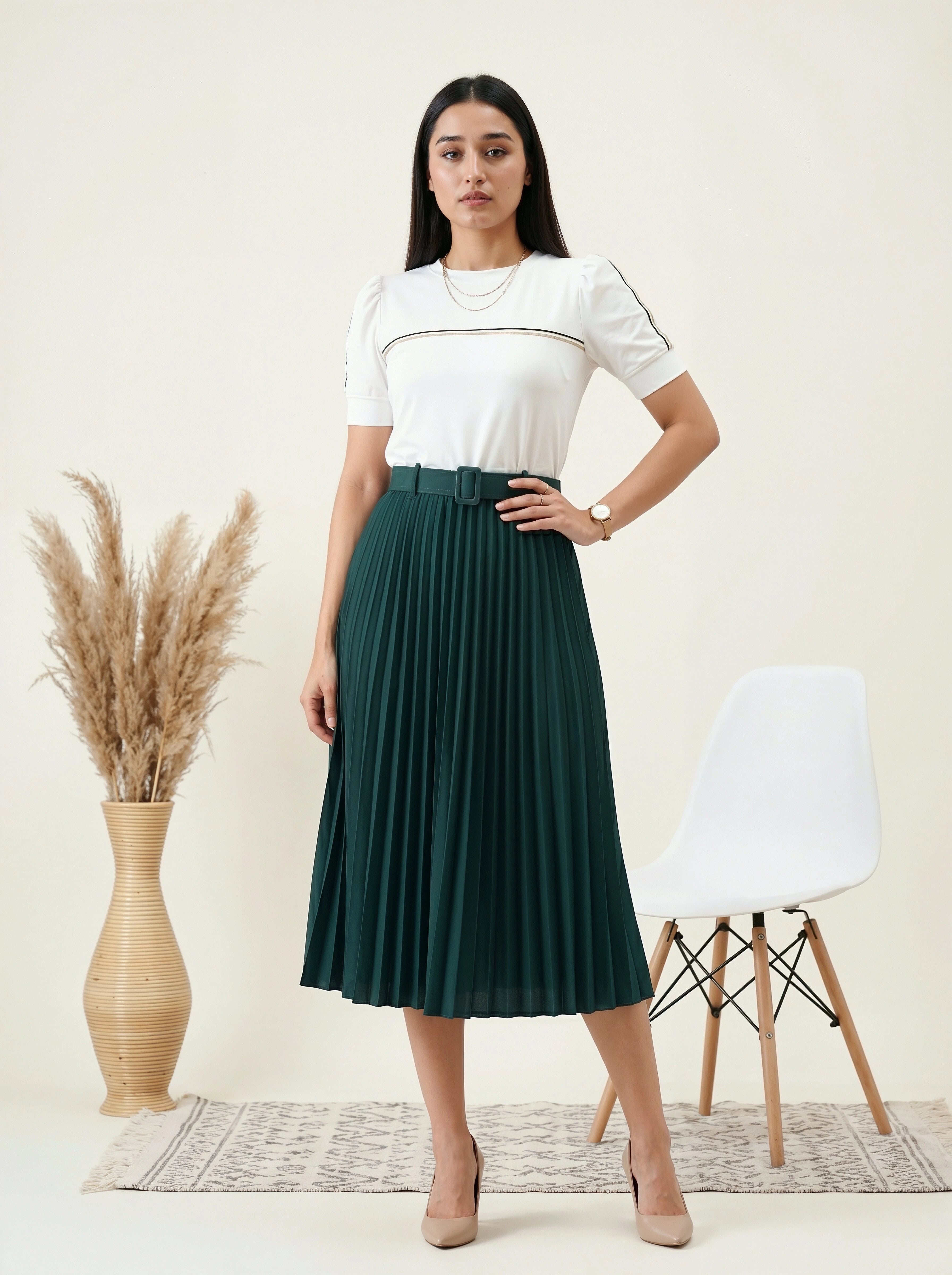Woman wearing a white top and green pleated skirt standing in a minimalistic room with a chair and plant.