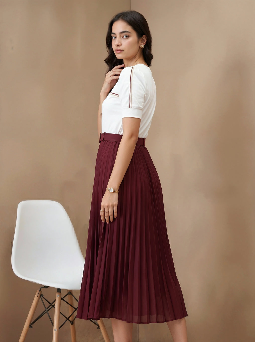 Woman wearing a white top and maroon skirt standing against a beige background with a white chair.