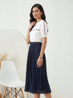 Woman wearing a white top with navy pleated skirt in a minimalistic room.