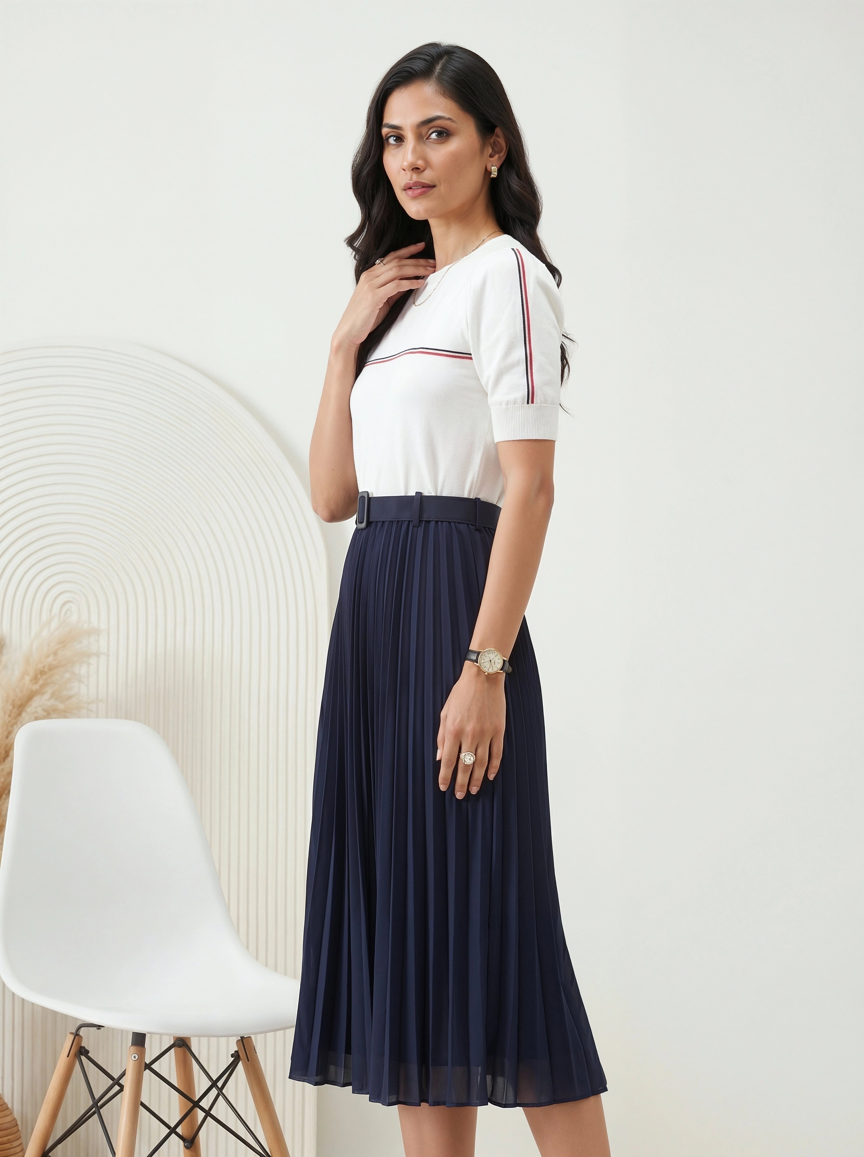 Woman wearing a white top with navy pleated skirt in a minimalistic room.