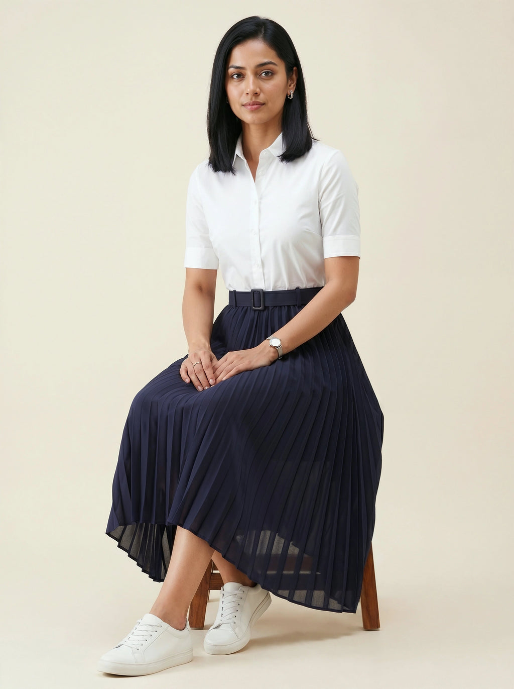 Woman wearing a white shirt and navy pleated skirt sitting on a stool against a beige background