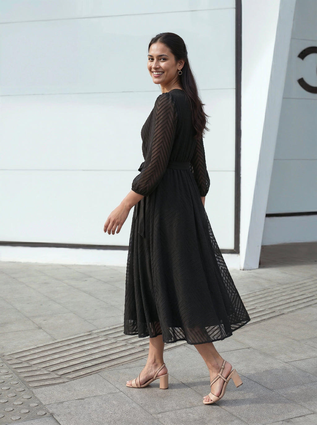 Woman wearing a black dress standing on a sidewalk with a light gray wall in the background