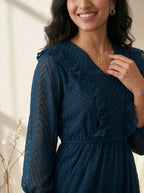 Woman wearing a dark blue dress with ruffled details, smiling indoors.