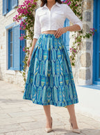 Woman wearing a white cropped shirt and blue patterned skirt standing in front of a stone building with blue shutters.