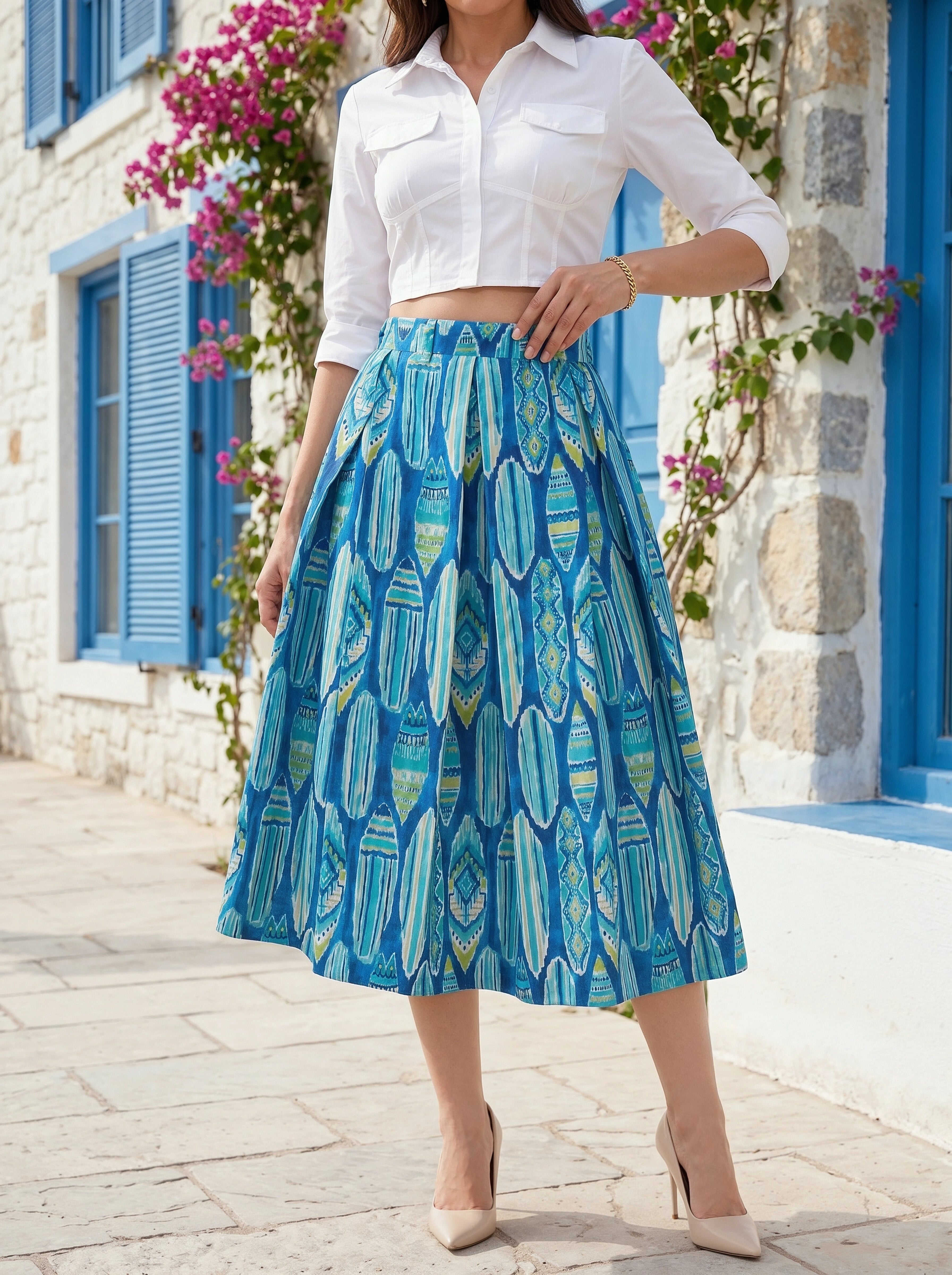 Woman wearing a white cropped shirt and blue patterned skirt standing in front of a stone building with blue shutters.