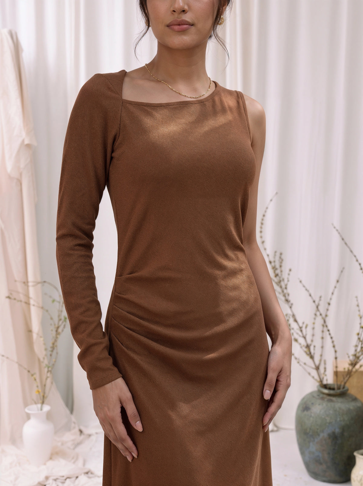 A woman wearing a long-sleeve brown knit dress with a square neckline and gathered side details, standing in a softly lit indoor setting with neutral decor.