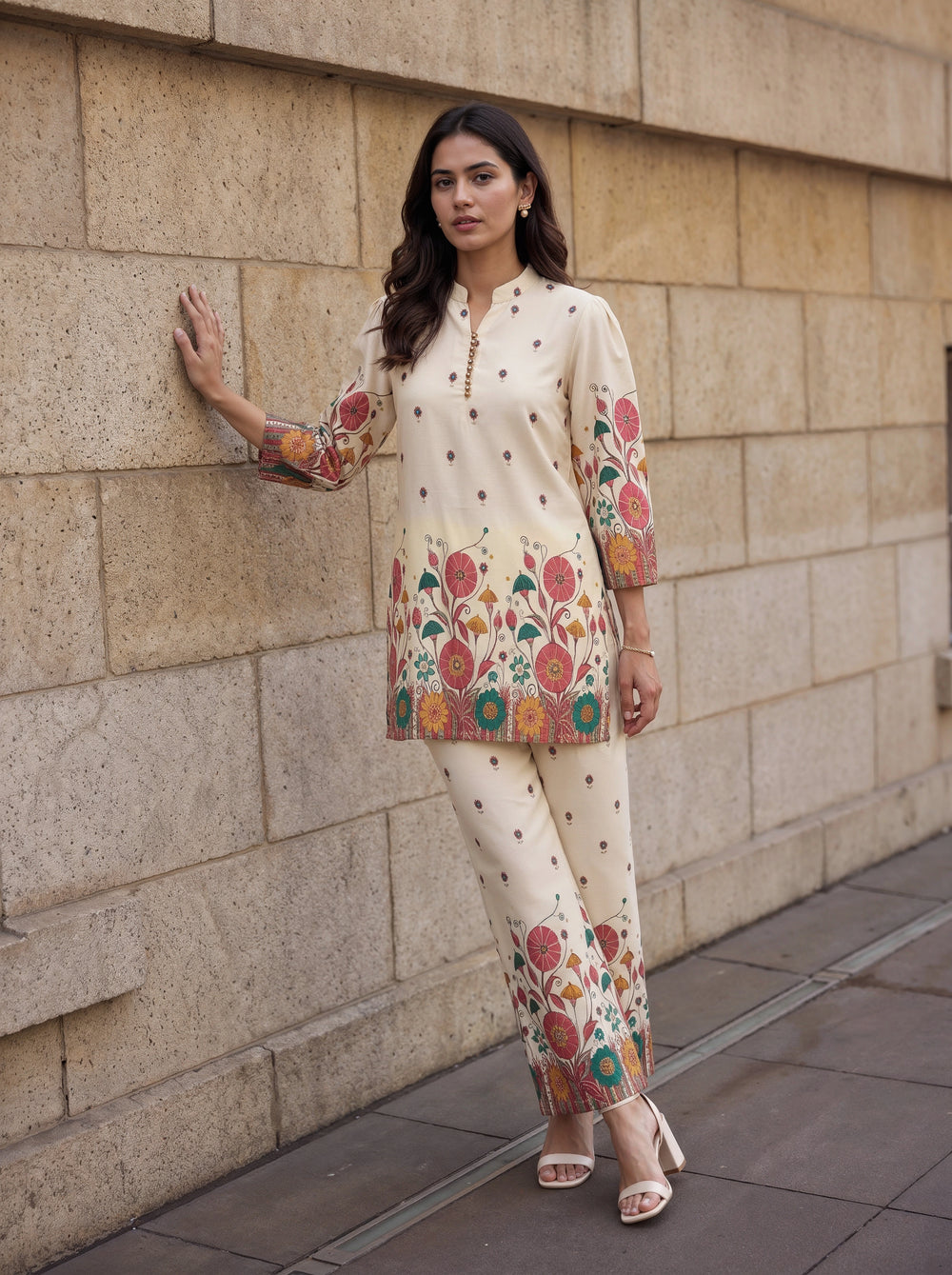 A woman models a cream-colored two-piece kurta and trouser set with colorful floral embroidery and small gold polka dots, standing against a beige stone wall.