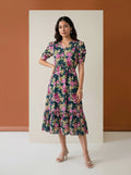 A woman modeling a short-sleeve midi dress with a dark teal floral print featuring pink, purple, and green flowers, standing in front of a minimalist studio backdrop with a terracotta-colored wall and a cream-colored panel.