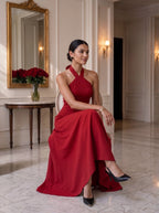 A woman wearing a floor-length red halter gown sits on a chair in an elegant indoor hallway with marble floors, a gold mirror, and wall sconces.