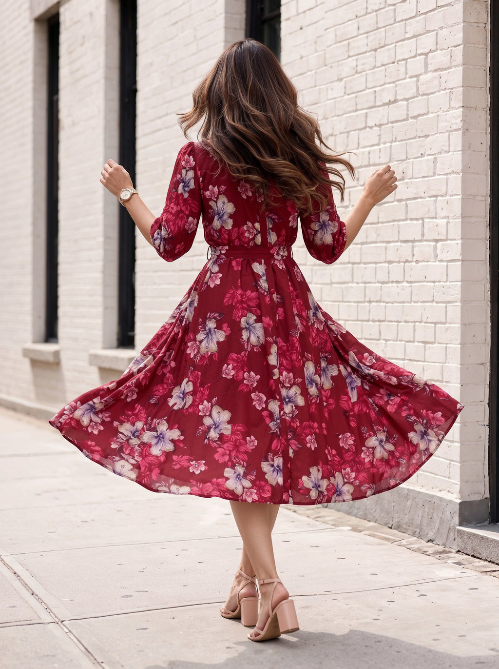 A woman wearing a red floral midi dress with three-quarter sleeves, shown from behind as she twirls on a sidewalk next to a white brick building.